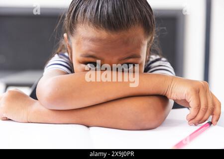 À l'école, une jeune étudiante biraciale se reposant la tête sur les bras dans la salle de classe, paraissant s'ennuyer Banque D'Images