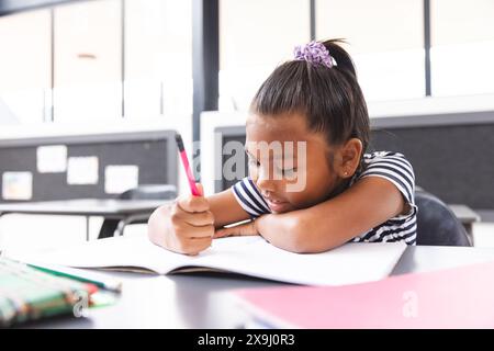 À l'école, en classe, jeune fille biraciale se concentrant sur l'écriture dans son cahier Banque D'Images