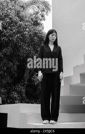 Une femme professionnelle en tenue d'affaires se tient en toute confiance à l'extérieur sur un escalier, encadrée par des arbres en arrière-plan Banque D'Images