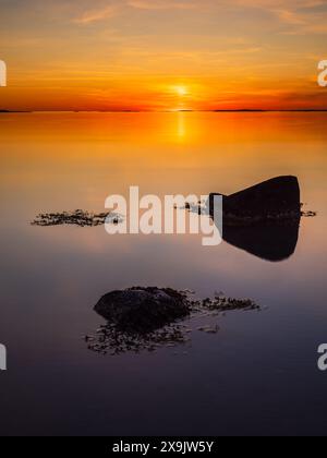 Un coucher de soleil serein sur la côte suédoise, avec deux grands rochers silhouettes partiellement submergés dans les eaux calmes et réfléchissantes. Le ciel est abla Banque D'Images