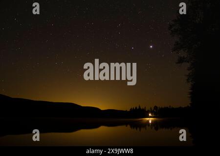 La lumière d'une cabane solitaire se reflète sur un lac tranquille sous un ciel rempli d'étoiles dans les bois du nord du Québec, Canada. Banque D'Images