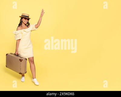 une femme robe inyellow et chapeau, portant une valise et prenant des photos par appareil photo vintage sur un fond jaune. Des gens heureux qui partent en vacances, vacat Banque D'Images