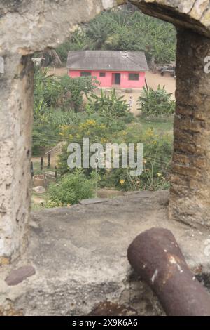 Cette image capture une scène vivante à travers une fenêtre voûtée à Fort Amsterdam, Ghana, surplombant un petit village avec une maison rose vif entourée de b. Banque D'Images
