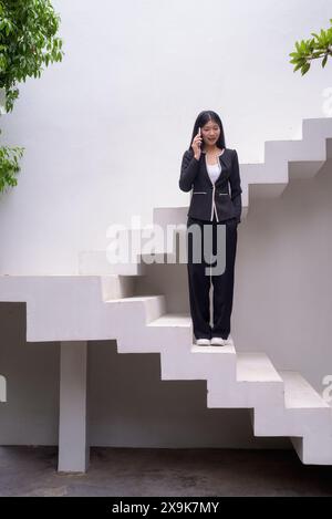 Portrait complet du corps d'une femme professionnelle en tenue d'affaires exsudant la confiance sur les étapes architecturales modernes, y compris la verdure et l'utilisation de pho mobile Banque D'Images