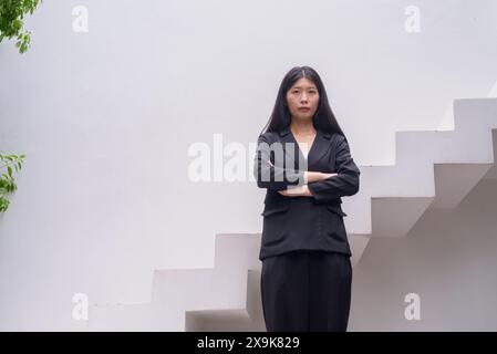 Portrait d'une femme professionnelle en tenue d'affaires exsudant la confiance sur les marches architecturales modernes, y compris la verdure avec les bras croisés Banque D'Images