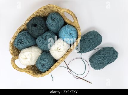 Photographie à plat d'une turquoise, vert foncé, sarcelle et boules de fil de laine blanche dans un panier avec des aiguilles circulaires sur un fond blanc Banque D'Images