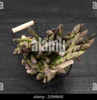 Gros plan d'asperges vertes luxuriantes nichées dans un panier métallique, directement après la récolte sur fond de bois noir Banque D'Images