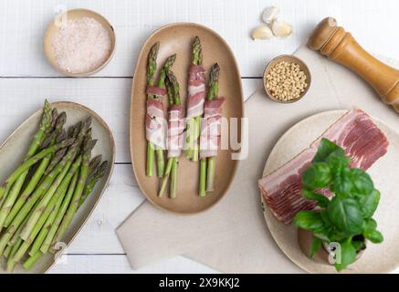 Asperges vertes croquantes montrées sur des assiettes beiges, prêtes pour le four. Encadré par des assiettes avec du bacon et d'autres ingrédients comme l'ail et les pignons de pin Banque D'Images