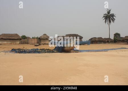 Un village de pêcheurs traditionnel avec des cabanes au toit de chaume et un bateau de pêche reposant sur la plage de sable. Les filets sont disposés pour sécher sous le soleil. Banque D'Images