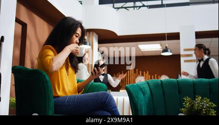 Touriste faisant défiler les applications de médias sociaux sur smartphone, en utilisant la connexion wi-fi gratuite dans le salon pour passer le temps avant de faire l'enregistrement. Femme asiatique se relaxant sur le canapé et planifier l'itinéraire du voyage. Banque D'Images