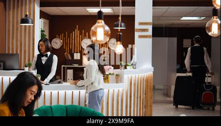 Le personnel de l'hôtel fournit des services de conciergerie de luxe pour les clients arrivant à la réception, aidant avec les sacs à roulettes et le processus d'enregistrement. Femme asiatique confirmant la réservation de chambre en ligne pour l'enregistrement. Banque D'Images