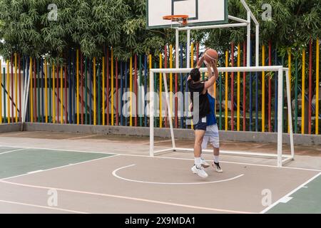 Deux hommes jouant au basket-ball sur un terrain extérieur, un sautant pour bloquer un tir. Poteaux verticaux colorés et feuillage vert en arrière-plan. Banque D'Images