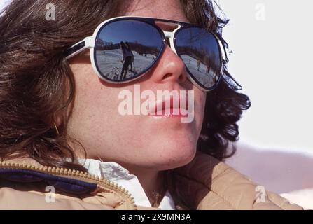 Gros plan de femme en lunettes de soleil aviateur reflétant les skieurs sur une pente enneigée, journée d'hiver lumineuse, portrait de style rétro avec scène de station alpine. Banque D'Images