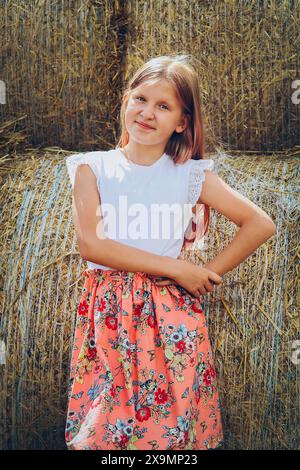 Une jeune fille se tient devant une balle de foin, portant une chemise blanche et une jupe florale, souriant dehors un jour d'été, Biélorussie, Minsk Banque D'Images