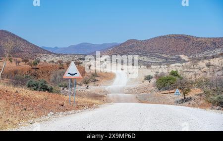 Route avec signalisation routière attention traversée de la rivière, piste de gravier mène à travers un paysage aride et sec avec des collines rouges et jaunes, Kaokoveld, Kunene, Namibie Banque D'Images