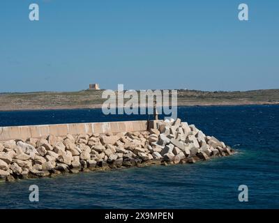 Un brise-lames de grandes pierres dans la mer avec une côte rocheuse en arrière-plan, l'île de Gozo avec des maisons historiques, des balcons colorés Banque D'Images