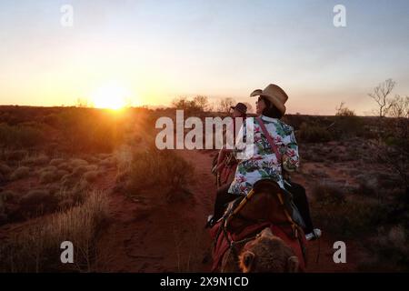 Deux femmes montent à dos de chameaux sur un sentier désertique dans l'outback australien jusqu'au coucher du soleil, les Olgas, Kata Tjuta à l'horizon lointain en Australie centrale Banque D'Images