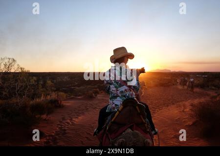 Une femme en promenade à dos de chameau avec un appareil photo photographie Kata Tjuta au coucher du soleil en Australie centrale sur un sentier de sable rouge dans le désert Banque D'Images