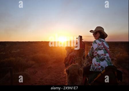 Une femme touriste sourit lors d'une balade à dos de chameau au coucher du soleil en Australie centrale avec le soleil sur Kata Tjuta, les Olgas et le large horizon désertique Banque D'Images