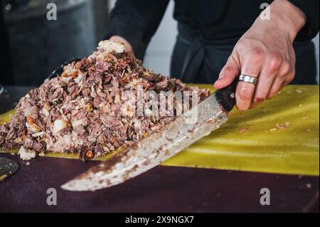 Les mains du chef masculin coupent la viande d'agneau bouillie avec un couteau pour cuisiner le Pilaf oriental traditionnel ouzbek n Centre de Pilaf d'Asie centrale Besh Qozon à Tachkent en Ouzbékistan Banque D'Images