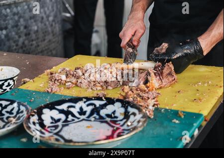 Le chef masculin coupe la viande bouillie d'un os d'agneau avec un couteau pour cuisiner le pilaf ouzbek traditionnel dans la cuisine d'un restaurant en Ouzbékistan Banque D'Images