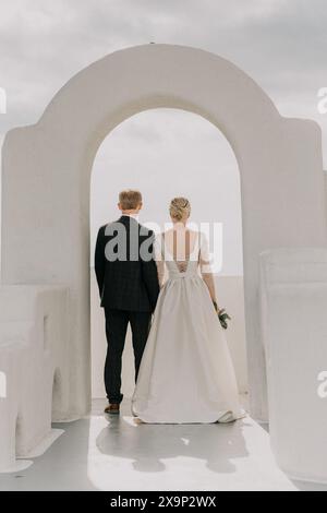 Une mariée et le marié marchent à travers une arche blanche par une journée ensoleillée. La mariée porte une robe blanche et le marié porte un costume. L'arche Banque D'Images