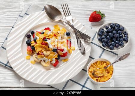 petit déjeuner sain de fraises fraîches, bleuets, bananes mélangées avec des flocons de maïs et yaourt sur plaque blanche sur table en bois blanc avec des ingrédients, Banque D'Images