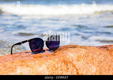 Une paire de lunettes de soleil élégantes reposant sur un grand rocher au bord de l'océan, reflétant les rayons du soleil. Le cadre suggère une journée de plage relaxante sous la chaleur Banque D'Images