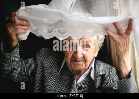 Une dame âgée regarde à travers un rideau de dentelle, ses yeux remplis d'histoires et de souvenirs. Banque D'Images