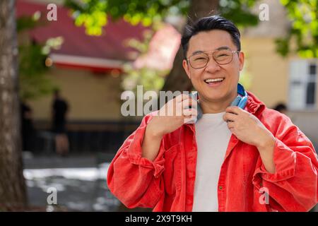 Portrait d'homme d'âge moyen asiatique souriant heureux écoutant de la musique décollant des écouteurs regardant la relaxation de l'appareil photo se sentir satisfait de bonnes nouvelles en plein air. Mec mûr chinois sur la rue de la ville urbaine. Horizontal Banque D'Images