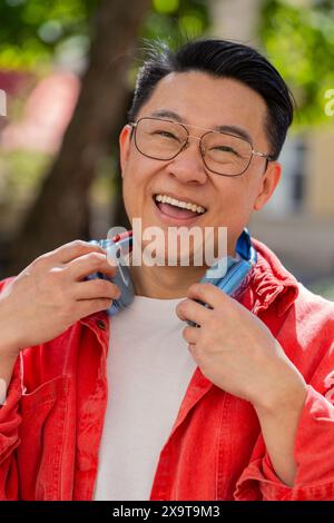 Portrait d'homme d'âge moyen asiatique souriant heureux écoutant de la musique décollant des écouteurs regardant la caméra, la détente se sentir satisfait de bonnes nouvelles en plein air. Mec mûr chinois sur la rue de la ville urbaine. Vertical Banque D'Images