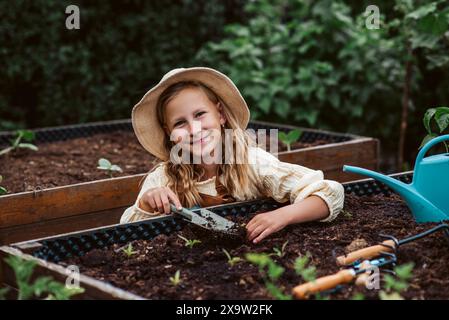 Fille prenant soin de petites plantes de légumes dans un lit surélevé, tenant une petite pelle. Enfance en plein air dans le jardin. Banque D'Images