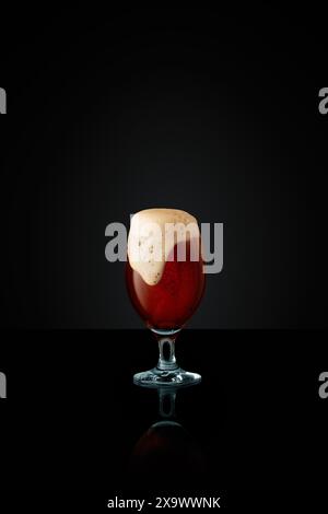 Gobelet en verre débordant de bière artisanale foncée, surmonté d'une mousse épaisse et crémeuse sur fond noir de studio. Banque D'Images