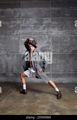 Danseur masculin, portant un manteau gris, un short blanc et un chapeau noir, se produit en studio avec un mur gris en arrière-plan. Homme en posture dynamique. Énergie et flexibili Banque D'Images