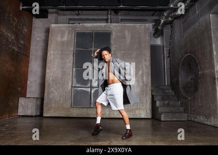 Danseur masculin, portant un manteau gris, un short blanc et un chapeau noir, se produit en studio avec un mur gris en arrière-plan. Homme en posture dynamique. Énergie et flexibili Banque D'Images