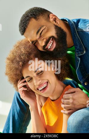 Un couple afro-américain heureux dans une tenue colorée sourit chaleureusement tout en embrassant sur un canapé jaune sur un fond gris. Banque D'Images