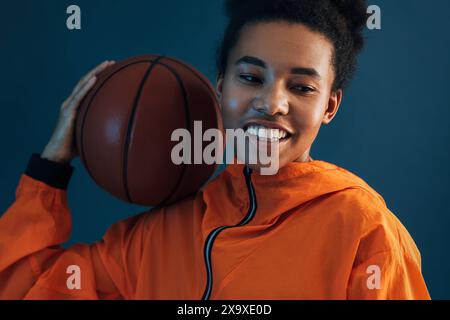 Gros plan d'une jeune joueuse professionnelle de basket-ball sur fond bleu. Femme joyeuse en vêtements de sport orange avec basket-ball sur son épaule. Fermer Banque D'Images