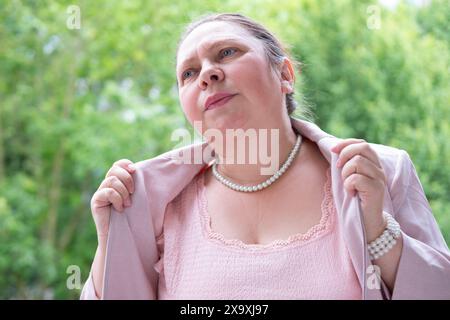 femme mature, 55 ans, élégamment habillée de rose se sentant malade, fan elle-même, vagues ménopausées, éprouver des bouffées de chaleur et de l'inconfort de la ménopause, fe Banque D'Images