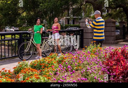 Un homme portant un haut aux couleurs vives prend une photo de deux jeunes femmes tenant leur vélo souriant et portant des robes très vives. Banque D'Images