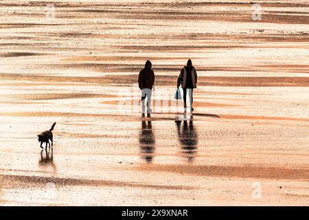 Deux personnes et un chien marchant dans la lumière qui s'estompe sur Fistral Beach à Cornwall. Banque D'Images