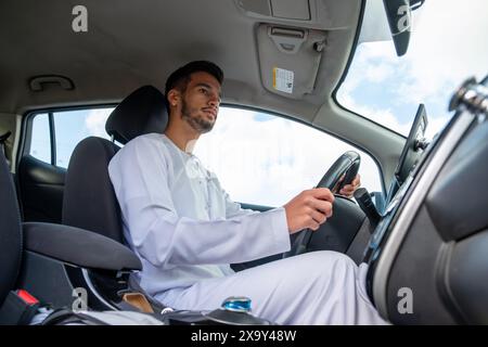 Un jeune homme joyeux avec une barbe, portant une tenue traditionnelle du moyen-Orient, sourit en toute confiance en étant assis dans sa voiture. Il semble s'amuser Banque D'Images