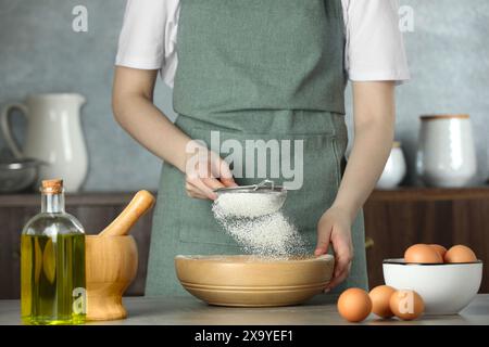 Femme tamisant la farine dans un bol à table dans la cuisine, gros plan Banque D'Images