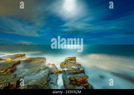 Une plage pittoresque la nuit avec des rochers, de l'eau, la pleine lune, et des nuages comme la lumière venant du ciel Banque D'Images