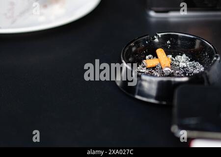 Gros plan d'un cendrier noir avec des mégots de cigarettes et des cendres sur une surface sombre. Banque D'Images