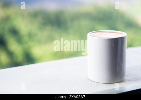 Une tasse en céramique blanche remplie de café placée sur un rebord à côté d'un cendrier noir avec une cigarette allumée. L'arrière-plan affiche une vue floue de vert hi Banque D'Images