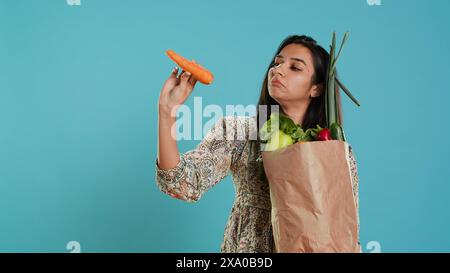 Femme avec un sac en papier dans les mains rempli de légumes vérifiant la carotte pour les impuretés. Client vivant conscient avec des produits d'épicerie achetés à utiliser comme ingrédients de cuisine, fond de studio, caméra A. Banque D'Images