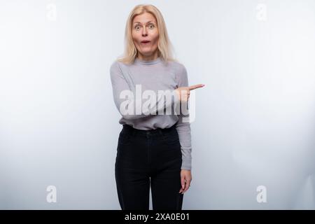 Mignonne femme blonde mature vêtue d'un col roulé gris pointe son doigt sur le côté dans la surprise Banque D'Images