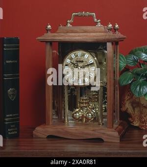 Horloge de cheminée en bois antique avec boîtier en verre et pendule en laiton sur la table polie contre le mur rouge, symbolisant le temps, la tradition, l'artisanat, la nostalgie Banque D'Images