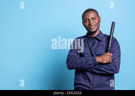 Agent de sécurité afro-américain se sent fier de son devoir en studio, posant avec confiance tenant son bâton vêtu d'un uniforme professionnel. Garde du corps habile debout sur fond bleu. Banque D'Images