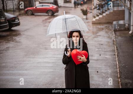 Une femme tenant un cœur rouge sous un parapluie clair. Le parapluie est ouvert et la femme est debout sous la pluie. Banque D'Images